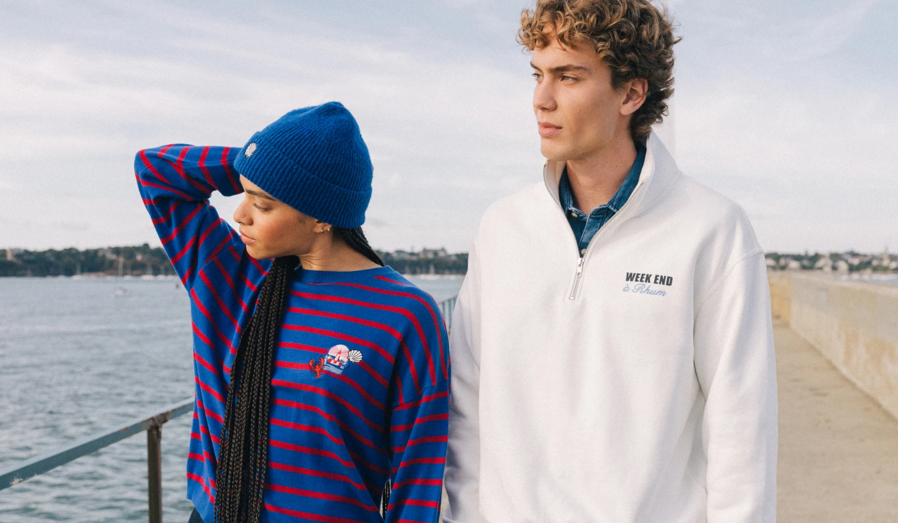 visuel mixte en bord de mer. La femme porte un pull rayé bleu et rouge. L'homme porte un sweat zippé blanc.
