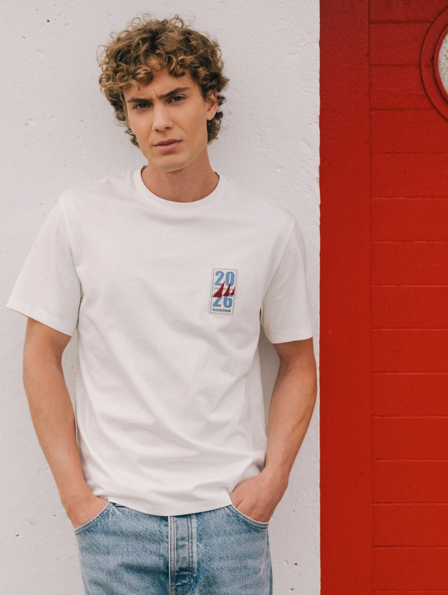 Visuel homme au pied du phare. Il porte un t-shirt blanc manches courtes avec un patch sur la poitrine.