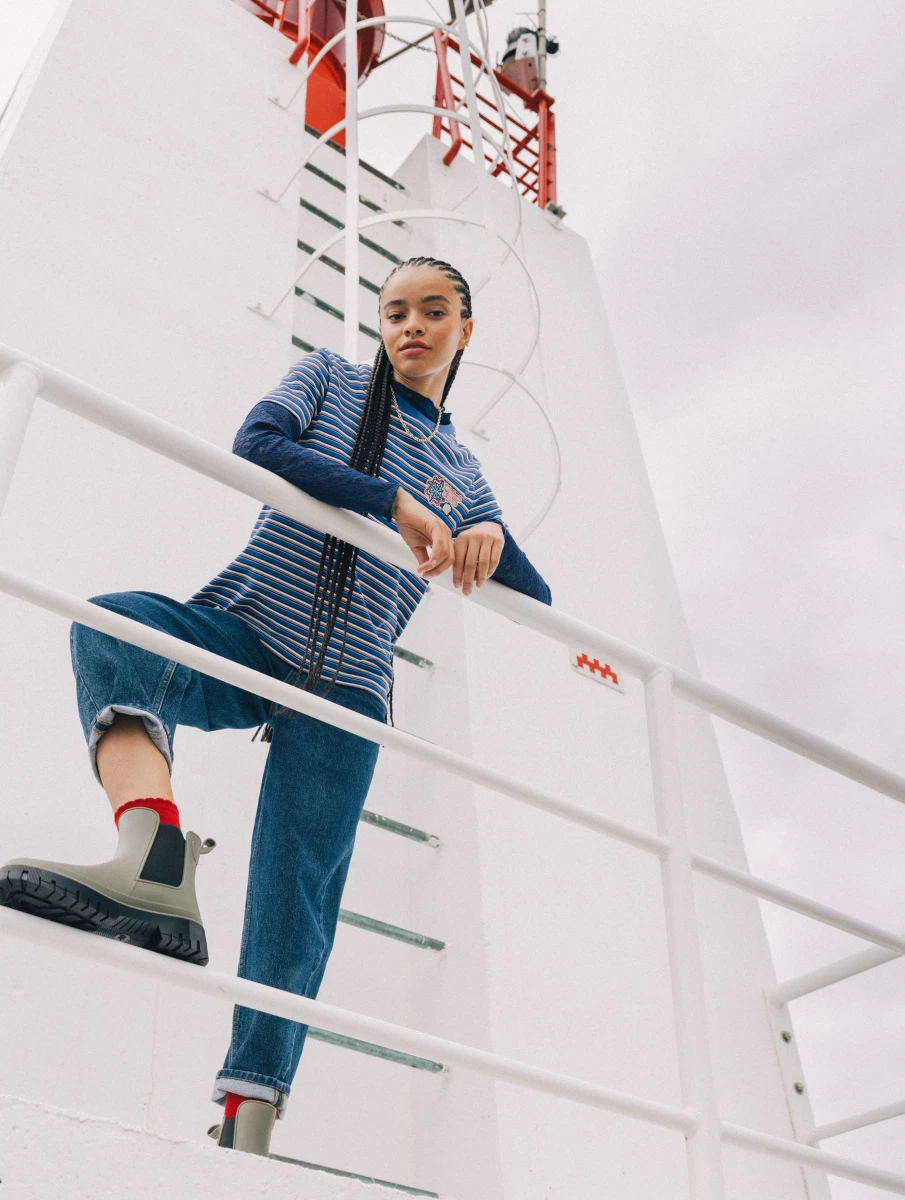 Visuel femme devant le phare. Elle porte un jean bleu et une marinière manches courtes par dessus un t-shirt bleu marine manches longues.
